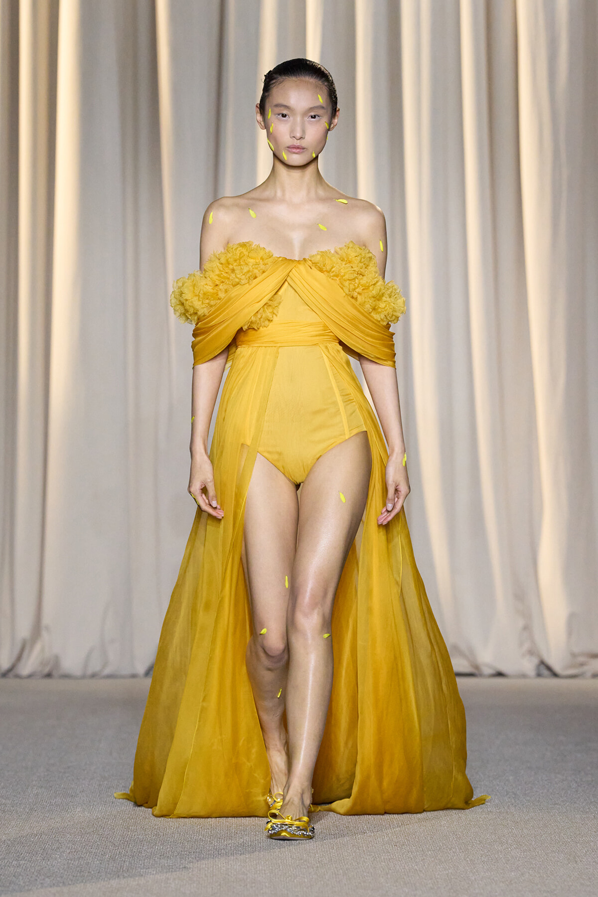 Model walking the runway in a yellow off-shoulder gown with a ruffled floral bodice and sheer cape on a neutral backdrop.
