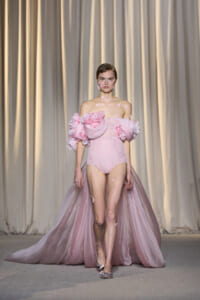 Model walking runway in an off-shoulder pink gown with voluminous floral sleeves and a long tulle cape, adorned with pink petals on face and shoulders.