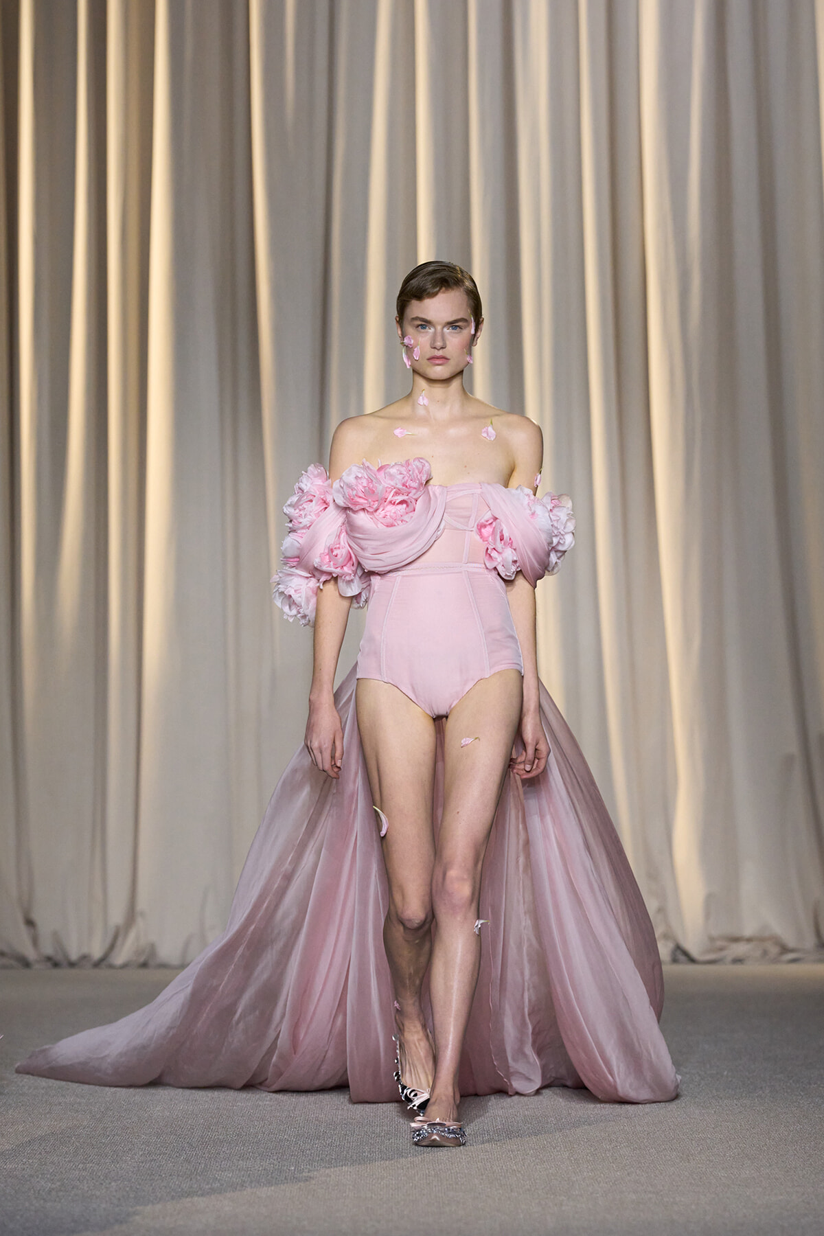 Model walking runway in an off-shoulder pink gown with voluminous floral sleeves and a long tulle cape, adorned with pink petals on face and shoulders.