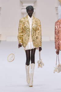 Model walking a runway in a gold sequin blazer, cream shirt, and pleated beige shorts, wearing cream lace-up knee-high boots and holding a small round beige handbag.