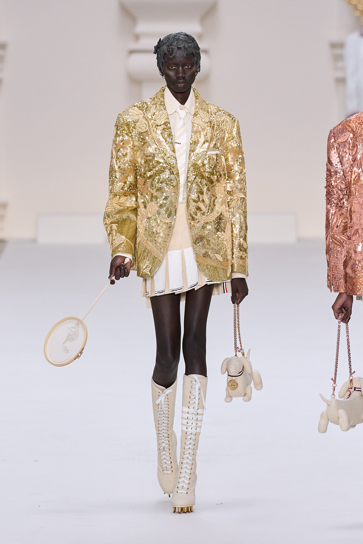 Model walking a runway in a gold sequin blazer, cream shirt, and pleated beige shorts, wearing cream lace-up knee-high boots and holding a small round beige handbag.