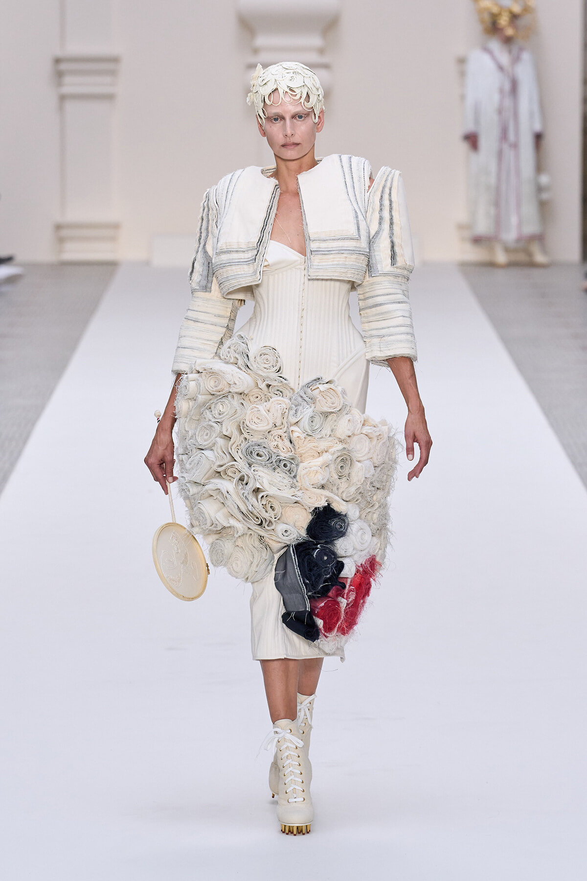 Model wearing an avant-garde white dress with layered rosettes, cropped jacket, headpiece, and lace-up boots on a runway, holding a circular clutch.