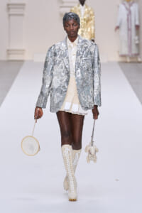 Model walks the runway in a silver sequined blazer, cream shirt and mini skirt, and cream lace-up boots, carrying a round embroidered bag and a plush bunny accessory.