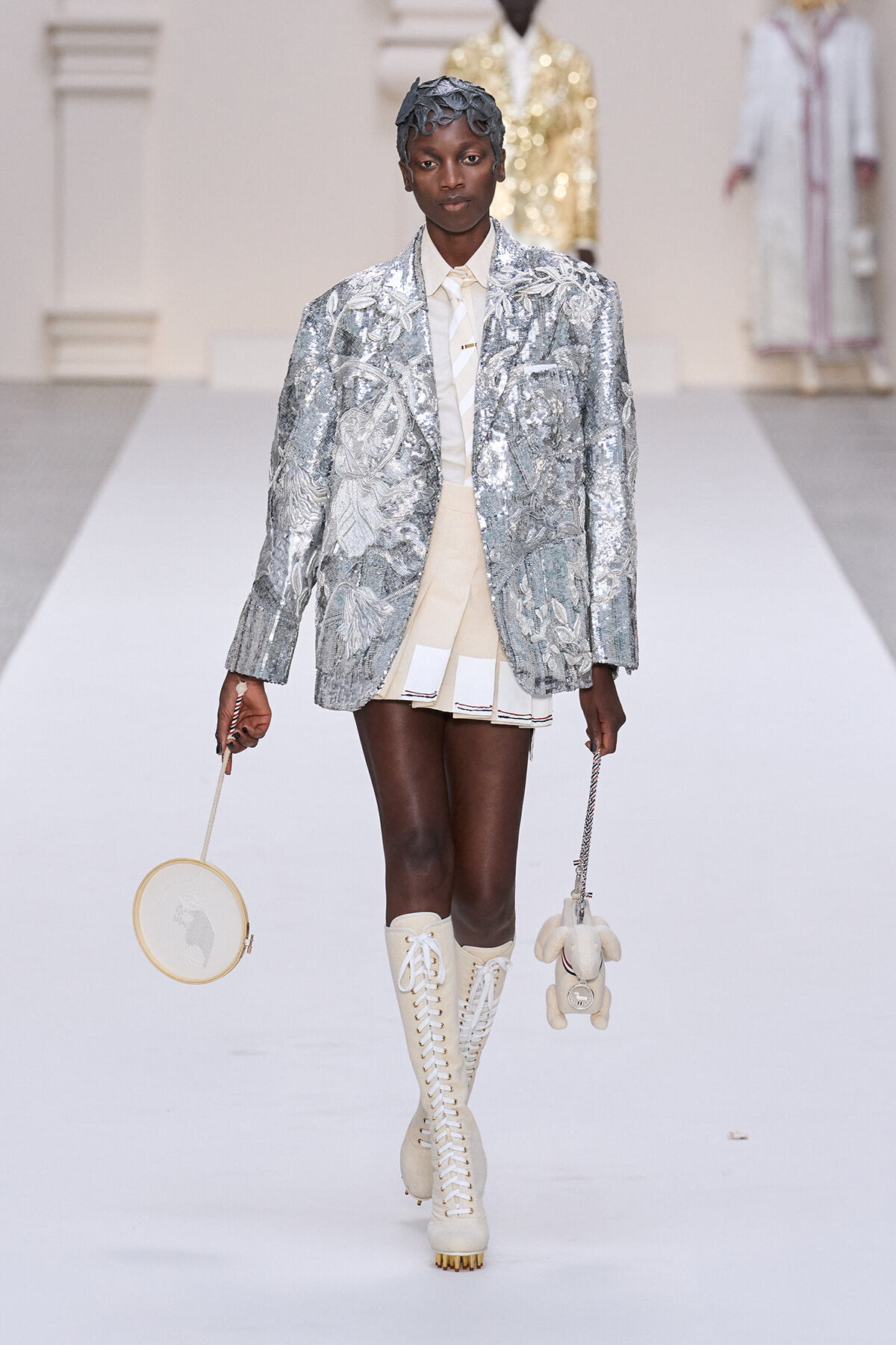 Model walks the runway in a silver sequined blazer, cream shirt and mini skirt, and cream lace-up boots, carrying a round embroidered bag and a plush bunny accessory.