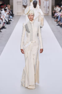 Model walks the runway in a cream-colored, tailored suit with patchwork panels and a circular headpiece.