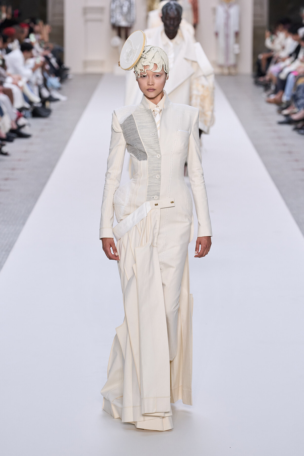Model walks the runway in a cream-colored, tailored suit with patchwork panels and a circular headpiece.