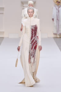 Runway model in a pale cream, high-neck gown with a bold red embroidered panel down the front and matching boots, walking a white catwalk.