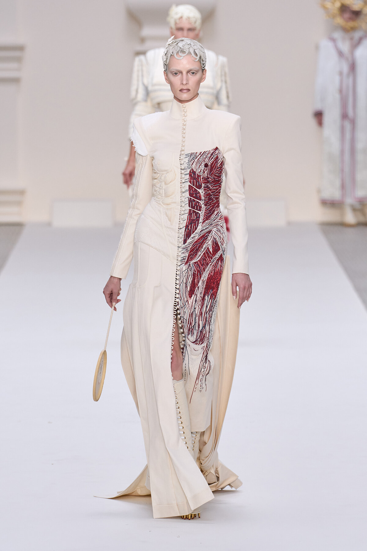 Runway model in a pale cream, high-neck gown with a bold red embroidered panel down the front and matching boots, walking a white catwalk.