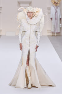 Model on a runway wearing an ivory, sculptural gown with oversized circular collar and intricate headpiece.