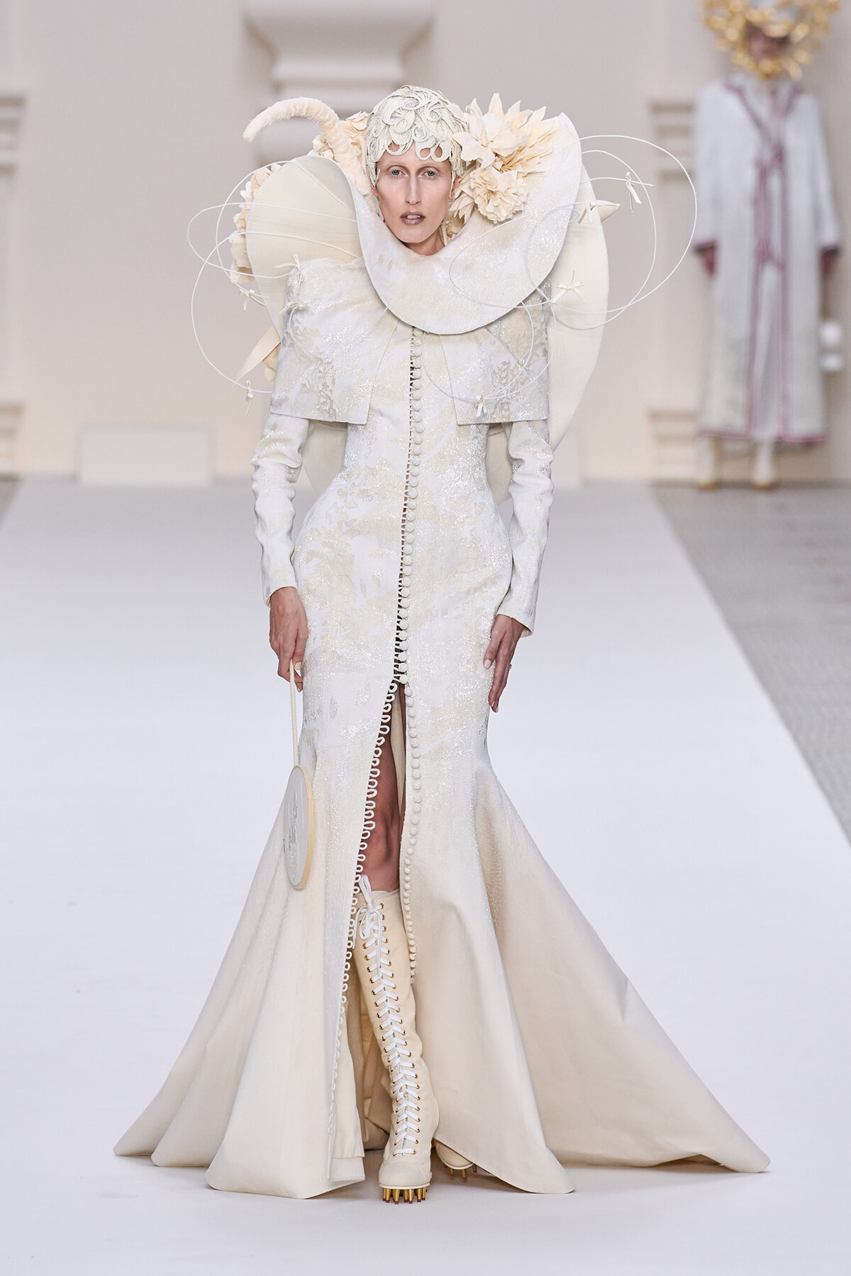 Model on a runway wearing an ivory, sculptural gown with oversized circular collar and intricate headpiece.