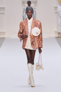 Model walks a runway in a rose-gold sequined blazer, cream shirt and pleated mini skirt with cream lace-up boots, carrying a round handheld accessory and a small dog-shaped bag on a pink chain.