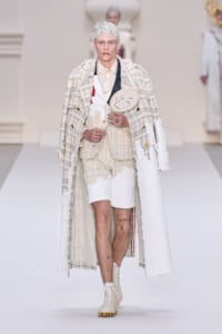 Male model walks the runway in a cream textured suit with a long white coat, holding a round fan with a smiling face.