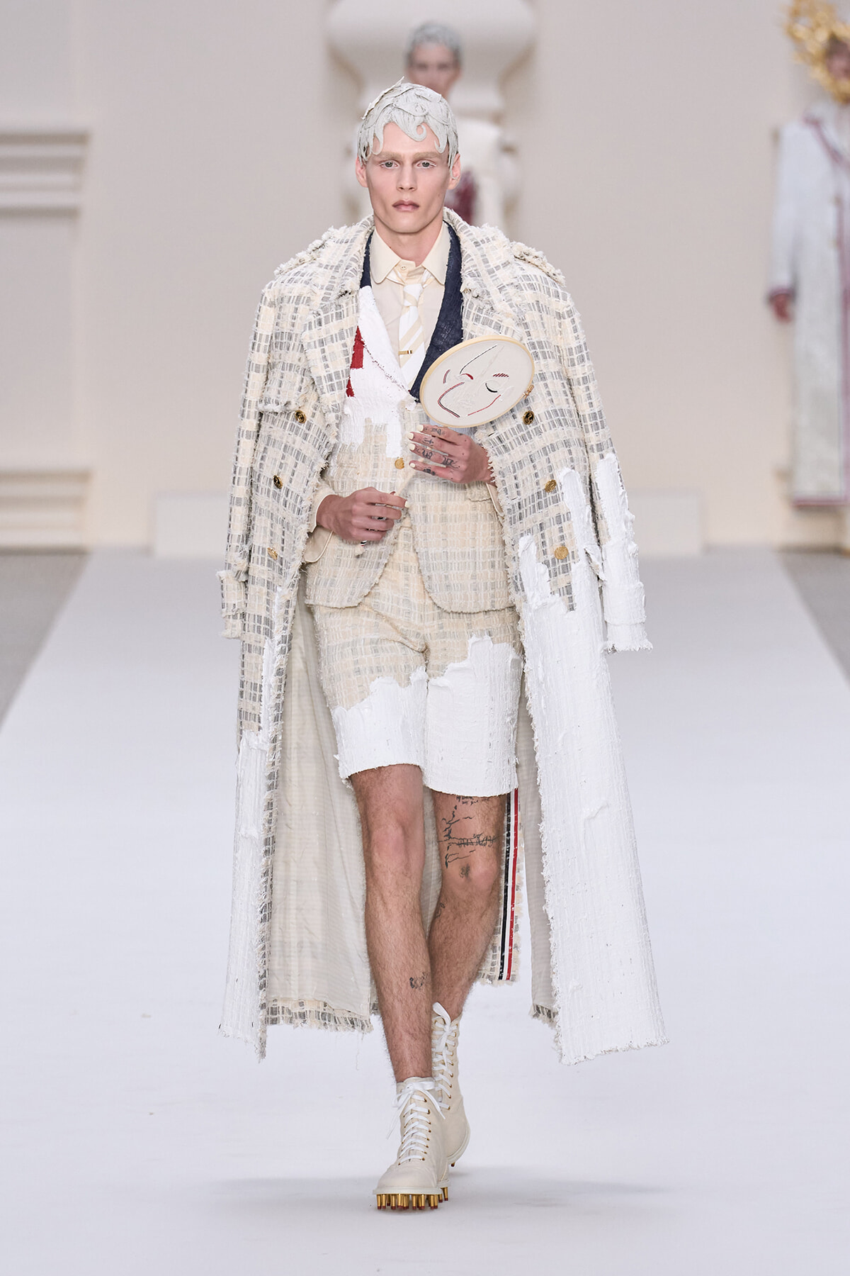 Male model walks the runway in a cream textured suit with a long white coat, holding a round fan with a smiling face.