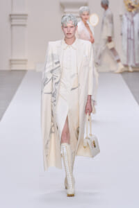 Model on a white runway wearing an ivory tailored suit with a long patterned cape, matching boots, and a cream handbag.