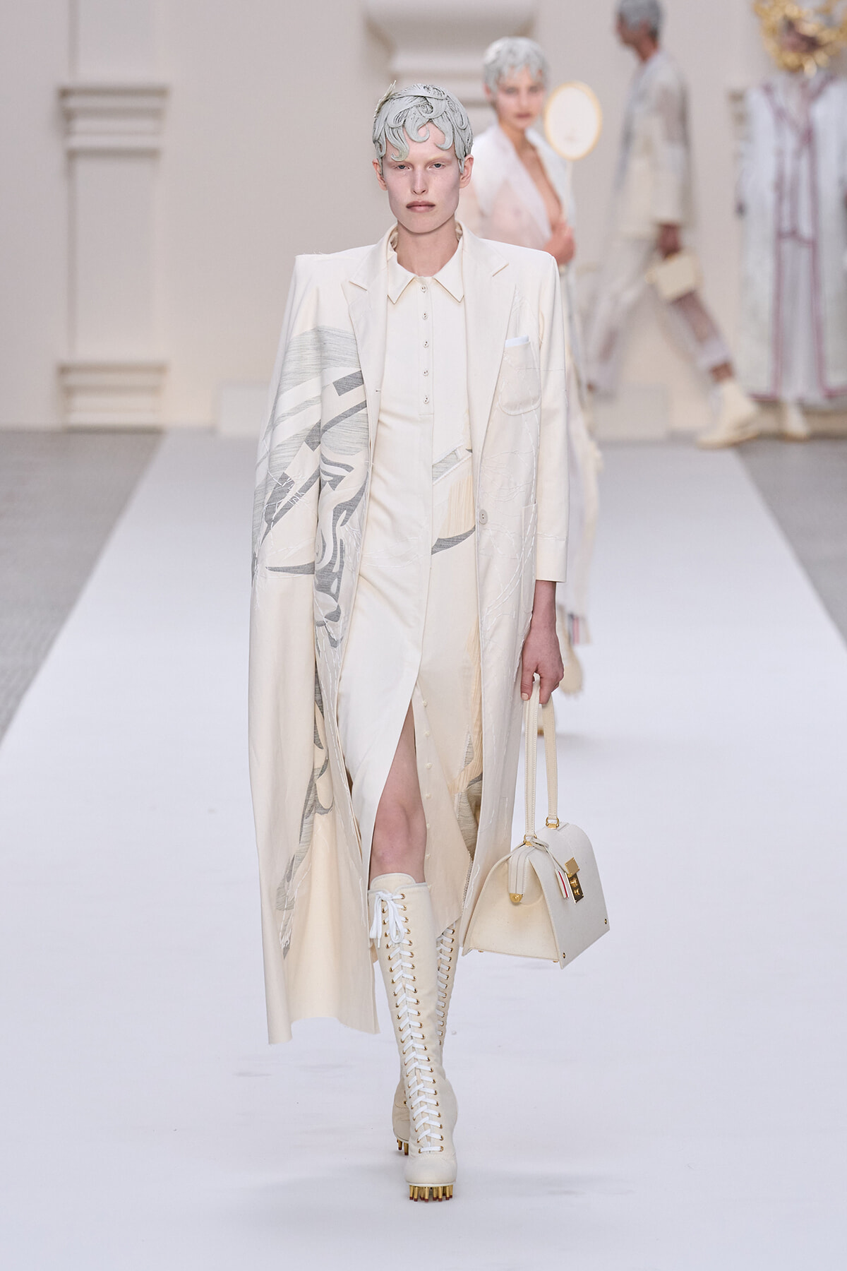 Model on a white runway wearing an ivory tailored suit with a long patterned cape, matching boots, and a cream handbag.