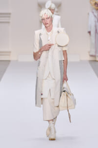 Male model walking a runway in an all-cream layered suit with grey panels, holding a round cream shoulder bag.