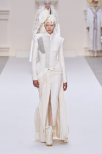 Runway model in an all-white sculptural suit with a gray panel, circular headpiece, and white lace-up boots walking a pale catwalk.