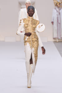 Model walking a runway in a cream and gold sequined suit with high collar, holding a round decorative fan. Gold embroidery and white thigh-high lace-up boots are visible on the avant-garde ensemble.