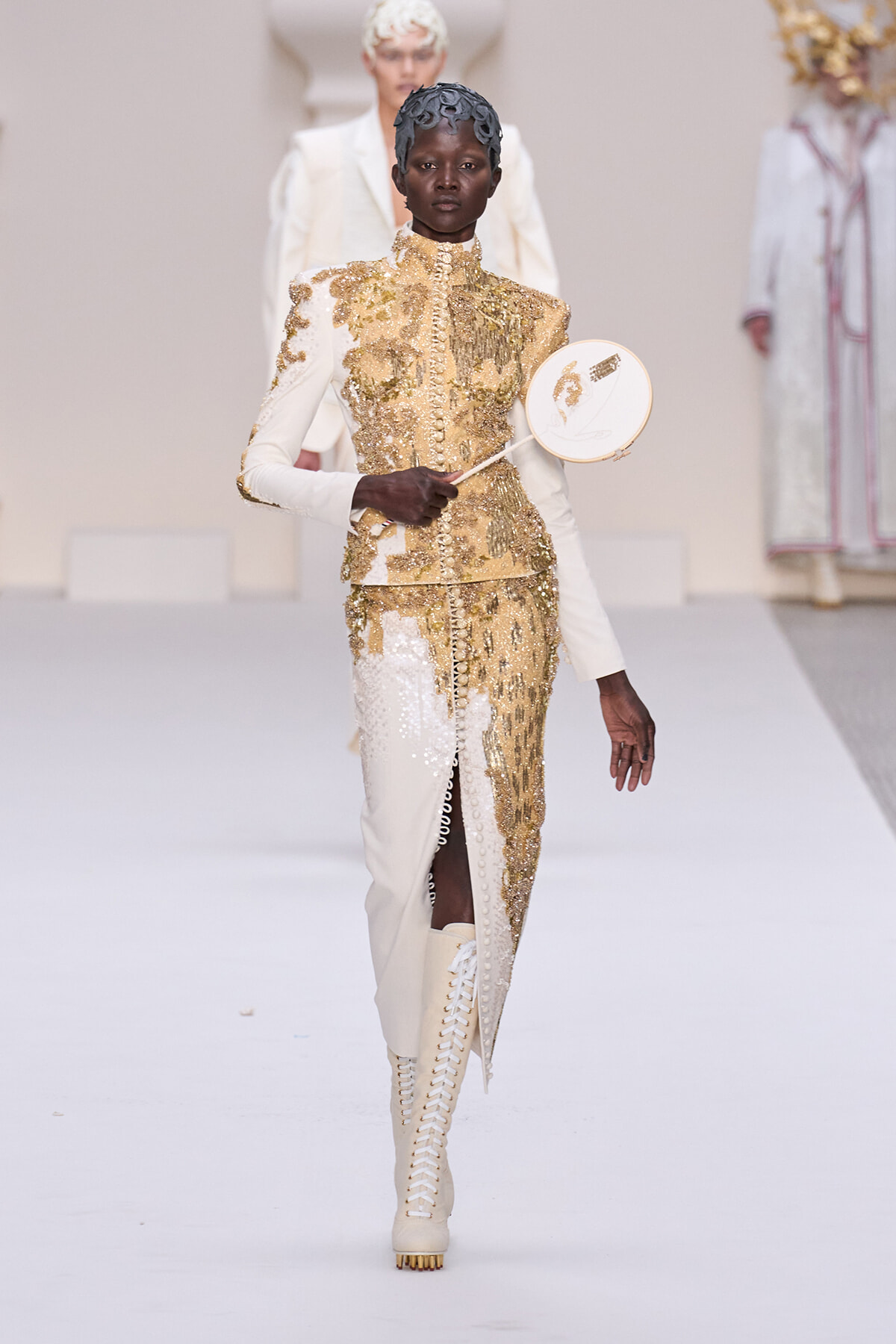 Model walking a runway in a cream and gold sequined suit with high collar, holding a round decorative fan. Gold embroidery and white thigh-high lace-up boots are visible on the avant-garde ensemble.