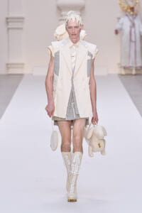 Male model walks the runway in a pale cream layered suit with gray panels, wearing tall lace-up boots and carrying a cream plush-dog bag and a round purse.