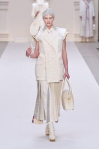 Runway model in an ivory layered suit with patchwork panels, holding a round handheld hat and a pale handbag.