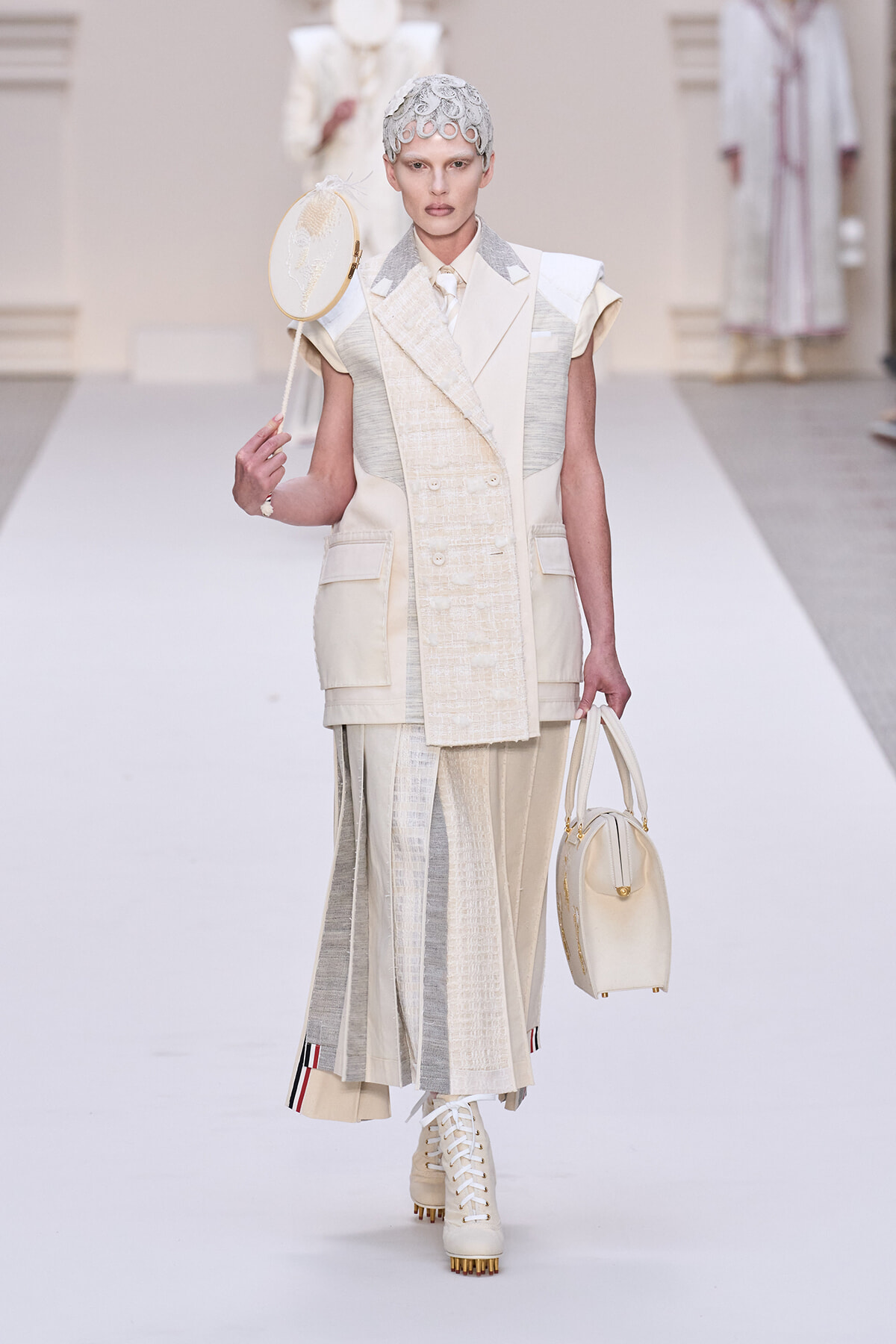 Runway model in an ivory layered suit with patchwork panels, holding a round handheld hat and a pale handbag.