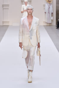 Male model in a white patchwork suit with sheer panels and a sculptural headpiece walks a runway, carrying a round white hoop bag and a small rectangular bag on a chain.