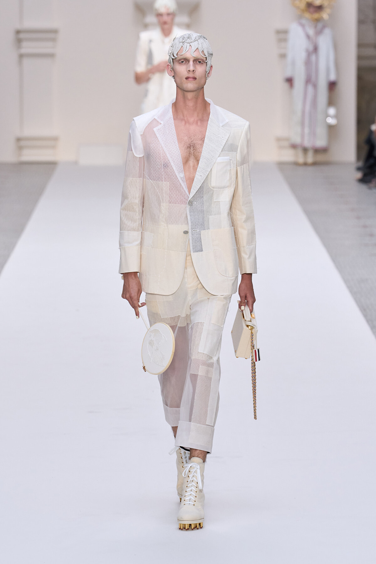 Male model in a white patchwork suit with sheer panels and a sculptural headpiece walks a runway, carrying a round white hoop bag and a small rectangular bag on a chain.