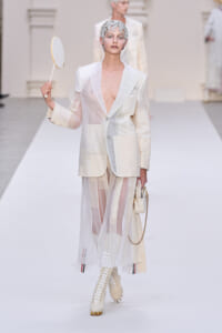 Model walking the runway in a cream patchwork suit with a sheer skirt, white lace-up boots, and a sculpted headpiece, holding a small circular mirror/lens prop.
