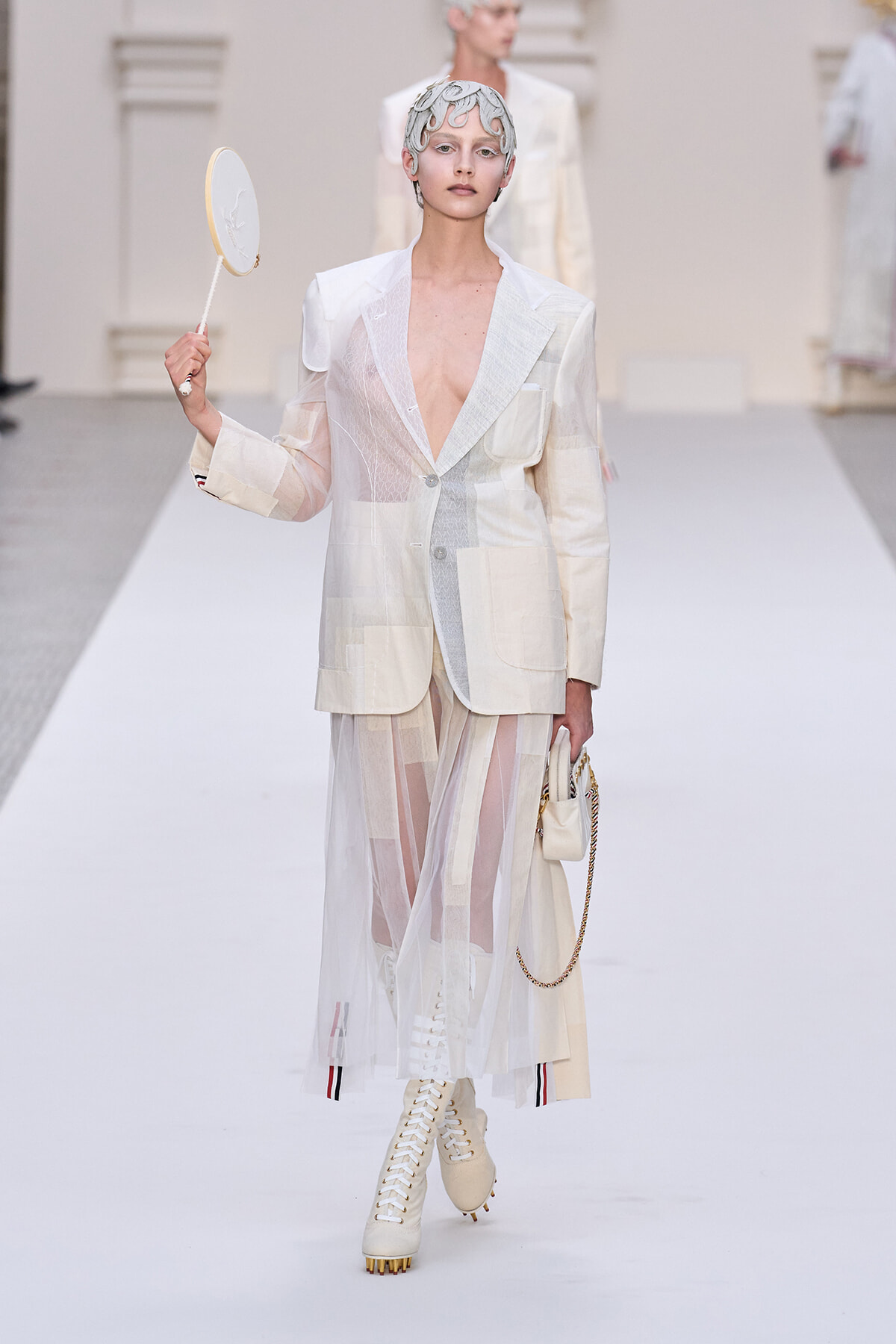 Model walking the runway in a cream patchwork suit with a sheer skirt, white lace-up boots, and a sculpted headpiece, holding a small circular mirror/lens prop.