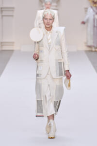 Model on a white runway wearing a cream patchwork suit with a headpiece, holding a circular mirror prop and a small cream bag.