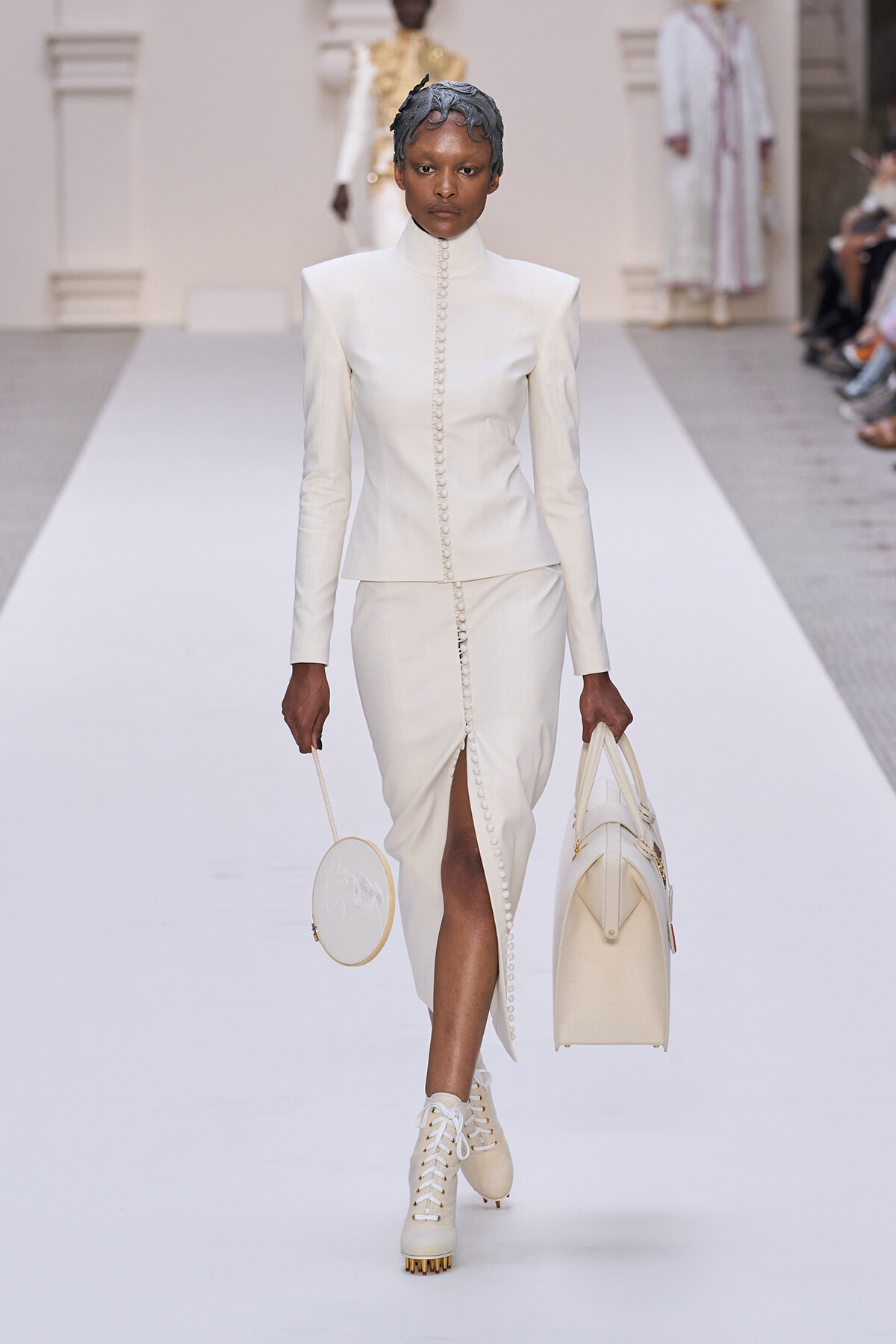 Model walks a runway in an ivory, tailored high-collar suit with a front button seam and a long skirt featuring a front slit; carries a round white purse and a large structured ivory bag, wearing white lace-up boots.
