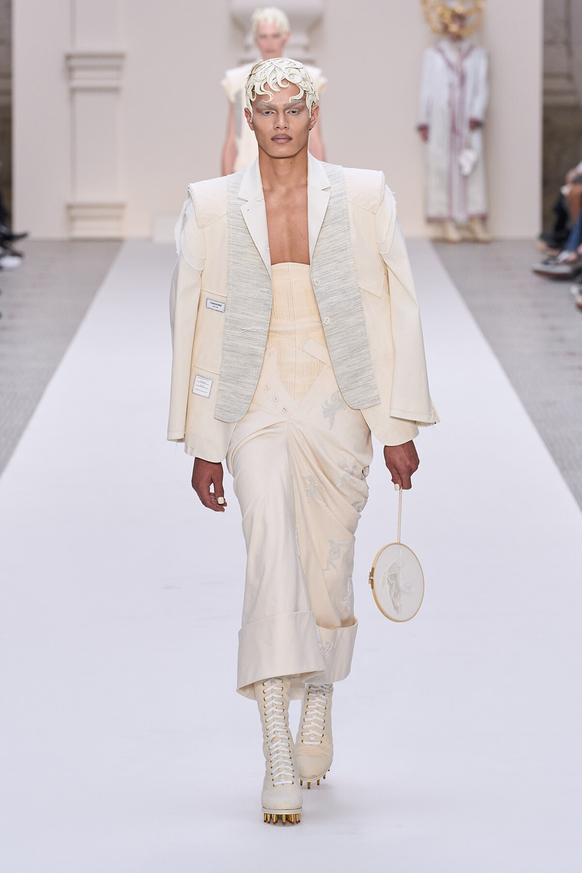 Runway look: ivory sculptural suit with oversized shoulders, corset-style top, embroidered skirt, lace-up boots, and a circular cream handbag.