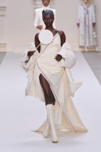 Black model walks the runway in an ivory draped gown with a white oversized coat, round handheld fan, and tall lace-up boots.