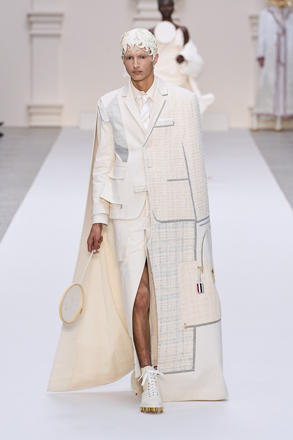 Male model walks a runway in an ivory, patchwork suit with a long textured cape and lace headpiece, holding a circular bag.