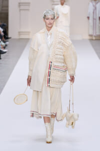 Model on a runway in an all-cream ensemble: oversized knit cardigan over a cream suit and pleated skirt, with lace-up boots and two bags as accessories.