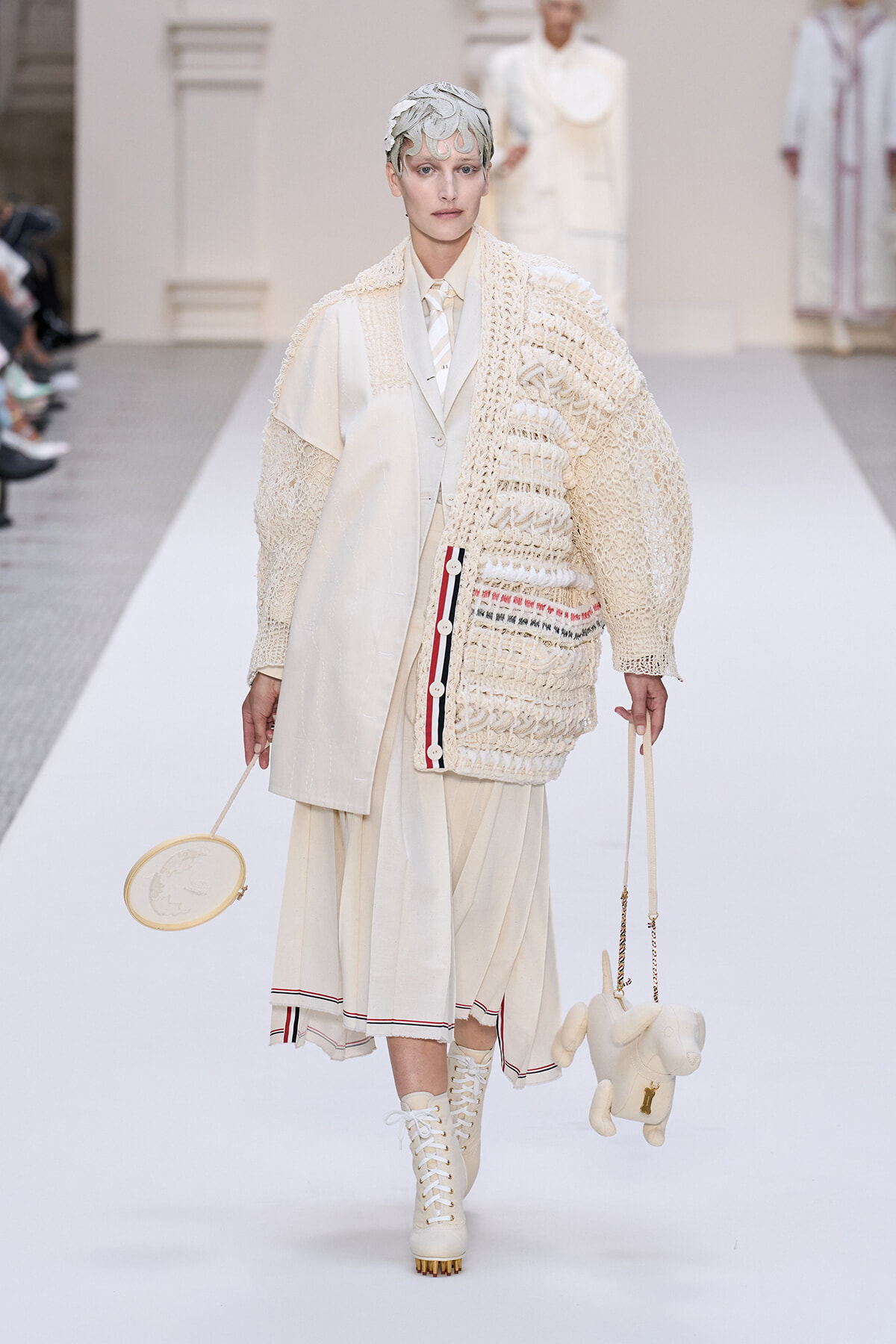 Model on a runway in an all-cream ensemble: oversized knit cardigan over a cream suit and pleated skirt, with lace-up boots and two bags as accessories.