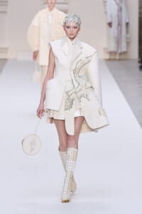 Model wearing an all-white avant-garde suit with sculpted cape detail, matching headpiece, knee-high lace-up boots, and a round handbag.