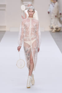 Model walking a runway in a sheer, embroidered beige dress with long sleeves, holding a circular embroidery hoop bag.
