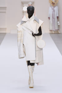 Model on a light runway wearing an oversized white trench with layered panels, gray shoulder details, a single textured sleeve, knee-high lace-up boots, and a round white handbag.
