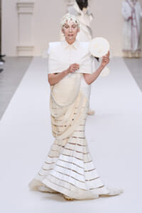 Model on a white runway wearing an all-white sculptural dress with a layered skirt and a decorative headpiece, holding a round fan.