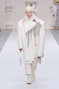 Model walking the runway in an oversized white coat with a draped textured panel, cream knit cap, and cream lace-up boots; holding a small circular bag.