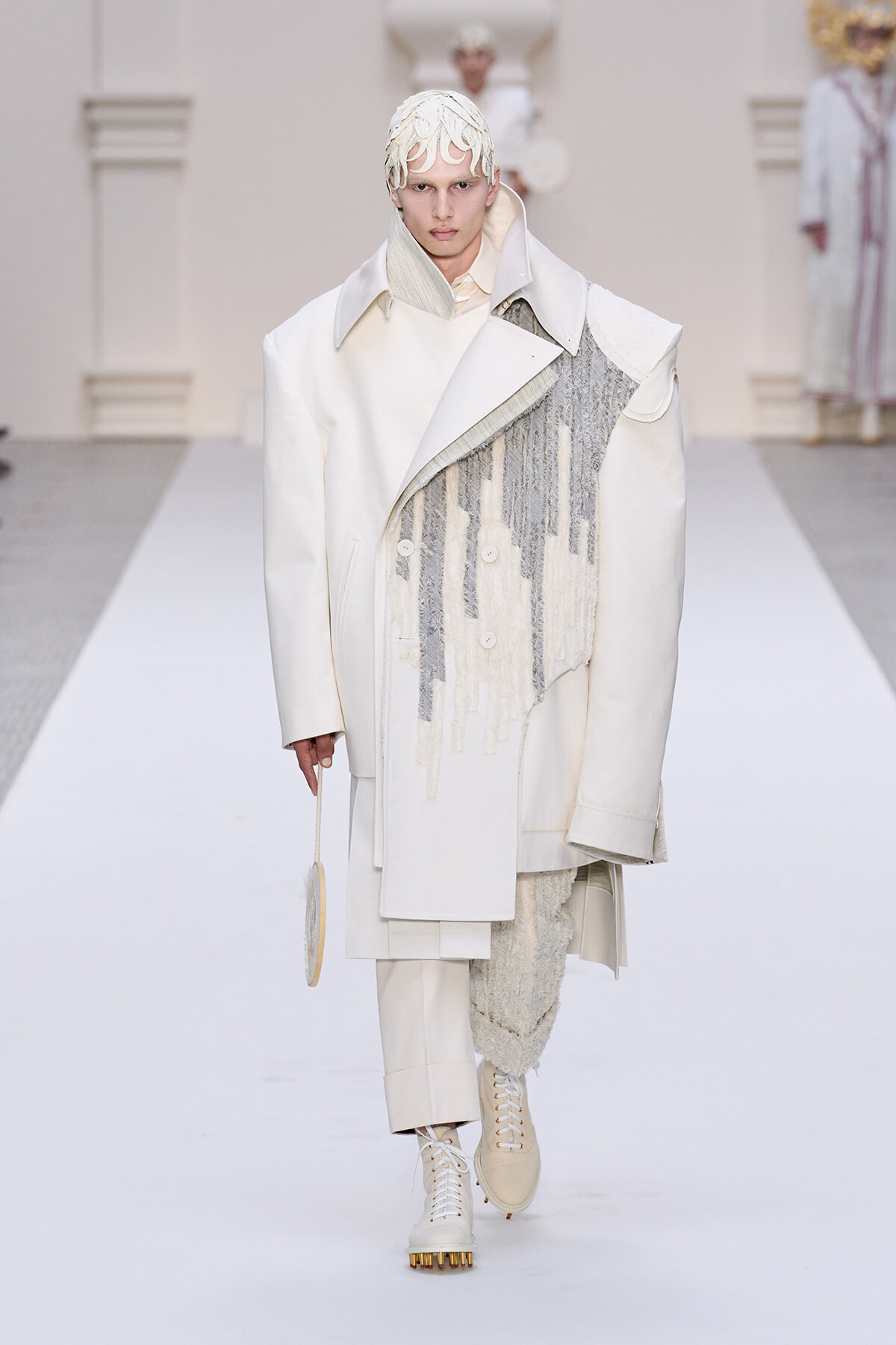 Model walking the runway in an oversized white coat with a draped textured panel, cream knit cap, and cream lace-up boots; holding a small circular bag.