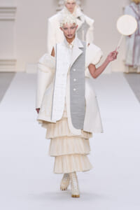 Model on a runway wearing an ivory asymmetric coat with a gray panel, a cream tiered skirt, lace headpiece, and a round mirror prop.