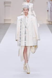 Model walking a runway in an oversized white coat with patchwork panels, cream outfit, and beige lace-up boots, wearing a metallic headpiece.