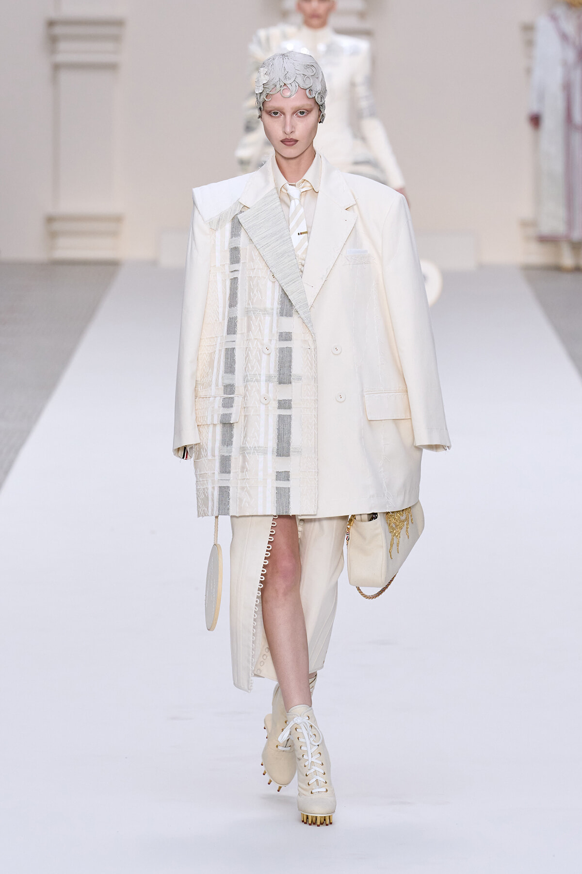 Model walking a runway in an oversized white coat with patchwork panels, cream outfit, and beige lace-up boots, wearing a metallic headpiece.