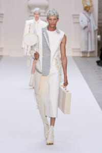Model on a white runway in a cream-toned ensemble with a gray panel, carrying a beige briefcase and a round cream prop in the other hand.