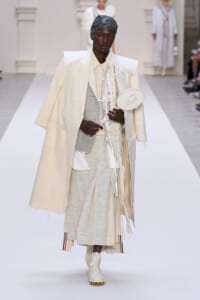 Male model on a white runway wearing an oversized cream cape over a pale gray suit, holding a circular white accessory.