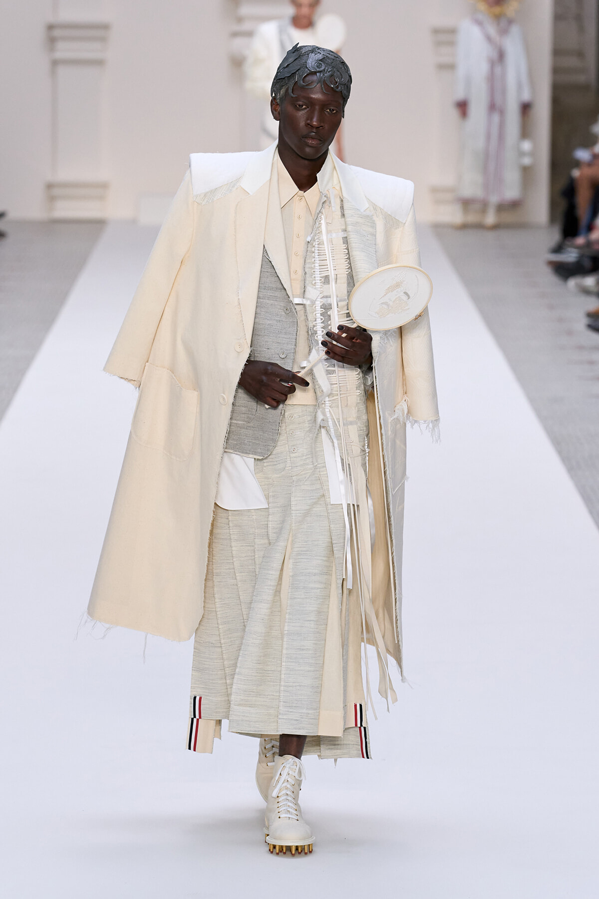 Male model on a white runway wearing an oversized cream cape over a pale gray suit, holding a circular white accessory.