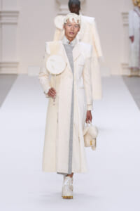 Model on a white runway wearing an ivory long coat with structured shoulders and a contrasting pale gray panel, paired with white boots and a sculptural headpiece; she carries a round white accessory with a face sketch and a small cream bear-shaped bag.